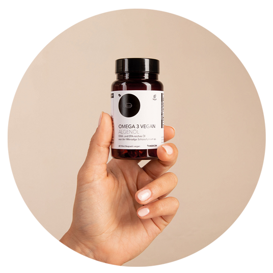 Eine Hand hält eine Flasche mit einem veganen Omega-3-Algenöl-Ergänzungsmittel vor einem hellbeigen Hintergrund.