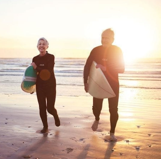 Zwei ältere Erwachsene in Neoprenanzügen tragen Surfbretter und gehen bei Sonnenuntergang lächelnd und glücklich am Strand entlang.