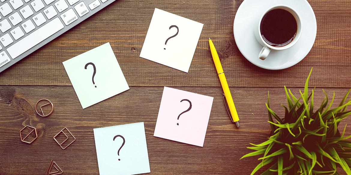 Vier Haftnotizen mit Fragezeichen, ein gelber Stift, Kaffee, eine Tastatur und eine Pflanze auf einem Holztisch.