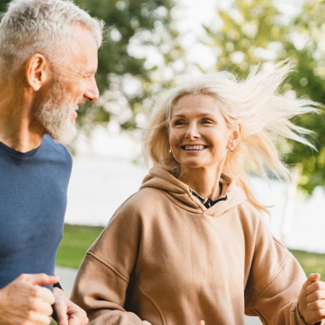 Älterer Mann und Frau joggen im Freien, lächeln sich gegenseitig an, mit Bäumen und Sonnenlicht im Hintergrund.