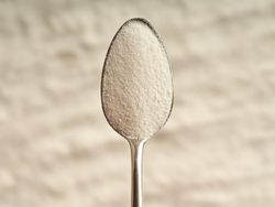 Nahaufnahme eines mit weißem Kristallzucker gefüllten Löffels vor einem unscharfen Hintergrund.