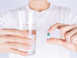Person, die in der einen Hand ein Glas Wasser und in der anderen Hand eine grün-weiße Kapselpille hält.
