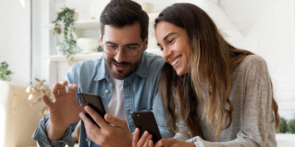 Ein lächelnder Mann und eine lächelnde Frau sitzen beieinander, schauen auf ihre Smartphones und wirken glücklich und beschäftigt.