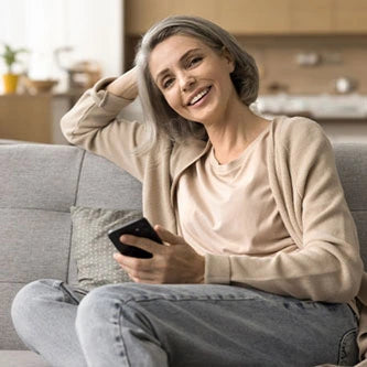 Lächelnde Frau mit grauem Haar, die auf einer Couch sitzt, ein Smartphone hält und in die Kamera schaut.