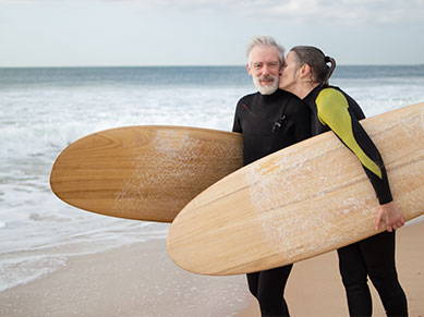 Ein älterer Mann und eine Frau mittleren Alters mit Neoprenanzügen und Surfbrettern am Strand.