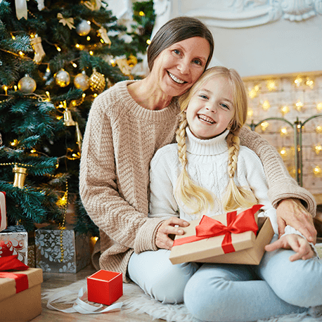 Eine Oma mit ihrer Enkelin sitzt vor einem Weihnachtsbaum und sie lächelln in die Kamera.
