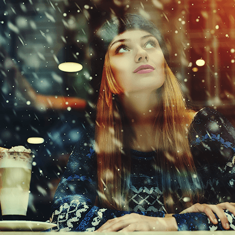 Eine Frau mit Pudelmütze sitzt in der Cafeteria und schaut auf die Schneeflocken draußen