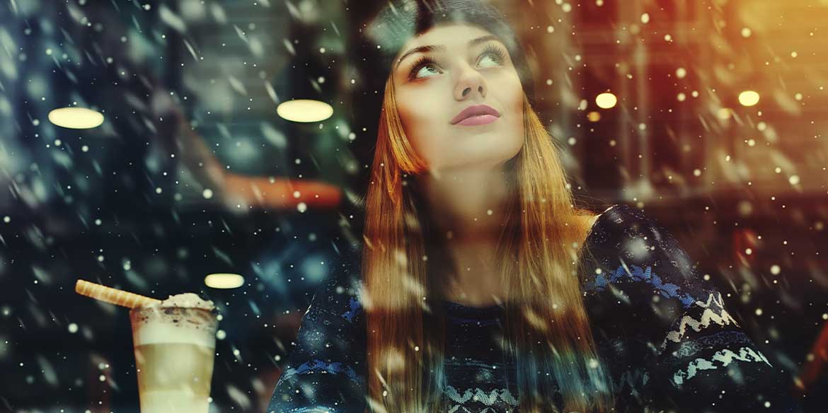 Eine Frau sitzt mit Pudelmütze im Café und schaut wie draußen die Schneeflocken fallen