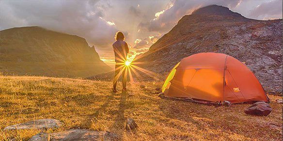 Eine Person erlebt einen Sonnenaufgang in einer Berglandschaft. Neben ihm steht ein Zelt.