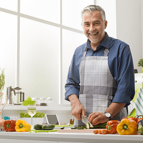 Ein Mann lacht in die Kamera. Er steht in der Küche und schneidet Gemüse.