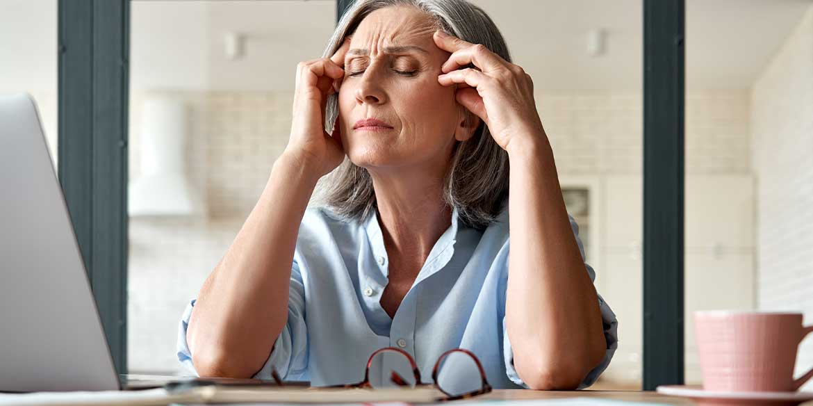 Eine ältere Frau mit geschlossenen Augen sitzt im Homeoffice und hält sich die Finger an die Schläfen