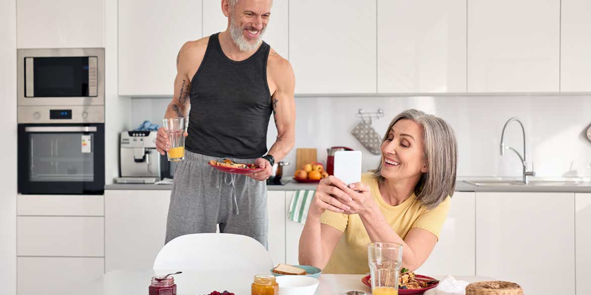 Ein älteres Paar in der Küche. Sie zeigt ihm lachend etwas auf dem Handydisplay