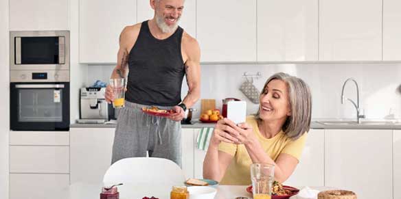 Ein älteres Paar in der Küche. Sie zeigt ihm etwas auf dem Handydisplay