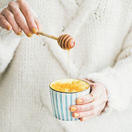Eine Frau mit gelb lackierten Fingernägeln und einem weißen Pulli süßt ihre Golden Milk (heiße Milch mit Curcuma) mit Honig