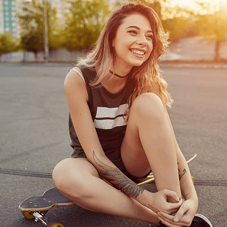 Junge Skaterin mit Tätowierungen sitzt auf einem Skateboard und lächelt