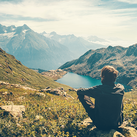 Ein entspannter Mann sitzt mit dem Rücken zur Kamera und schaut auf eine atemberaubende Berglandschaft mit See