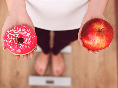 Foto von oben auf eine Frau, die barfuß auf der Waage steht und in der einen Hand einen Apfel, in der anderen einen Donut hält.